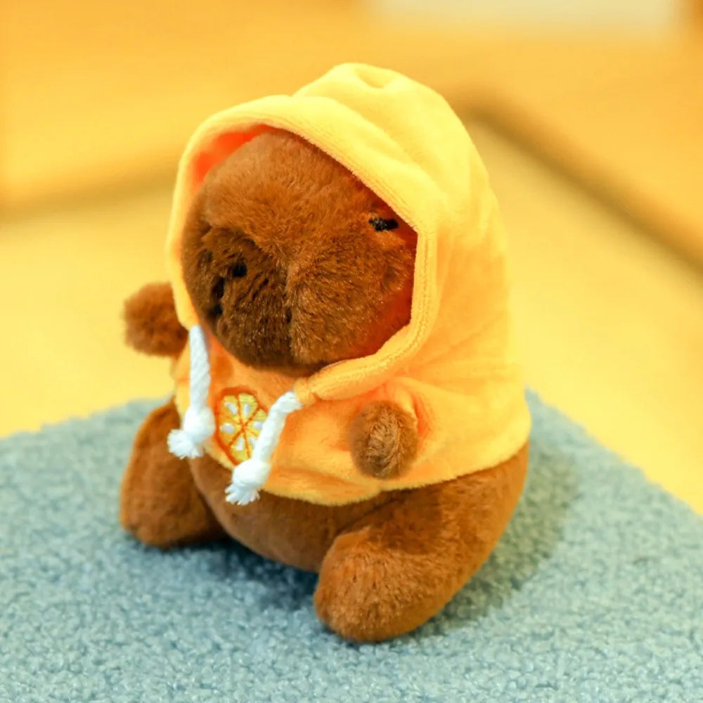 Peluche capybara brune en capuche jaune à motif citron, assise sur un tapis turquoise